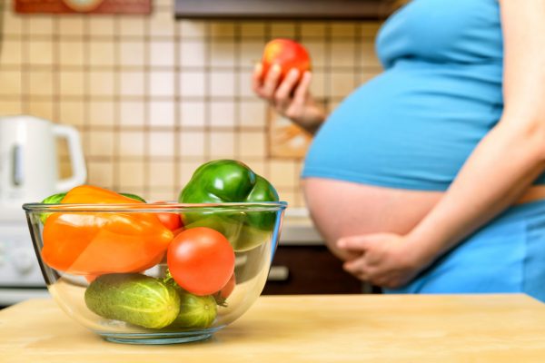 Овощи в прозрачной тарелке на столе, на заднем плане беременная женщина