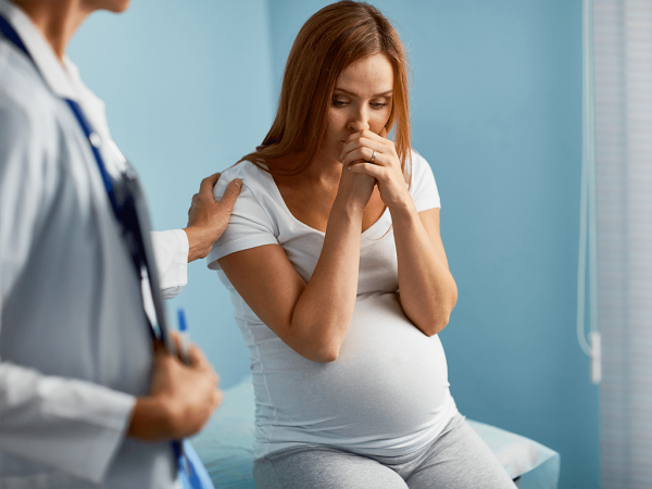 Беременная женщина прижимает руки к лицу, сидя на кушетке перед врачом