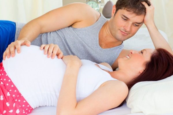 Супруги лежат на кровати, рука мужа находится на животе беременной жены