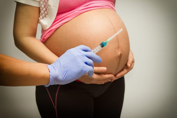 Медик держит в руке шприц, рядом стоит беременная женщина