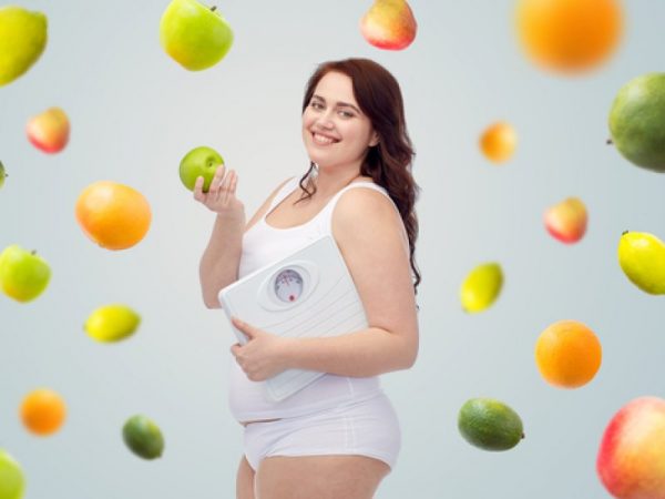 Фотография полной беременной женщины с яблоком и весами в руках
