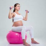 Беременная на фитболе выполняет упражнение с гантелями