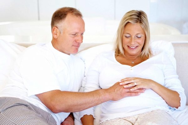 Мужчина с сединой и беременная женщина в возрасте сидят на диване и держат руки на беременном животе