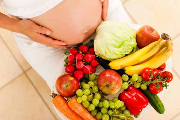 На коленях у беременной поднос со свежими овощами и фруктами