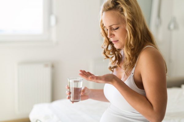 беременная держит в одной руке таблетку, в другой — стакан с водой